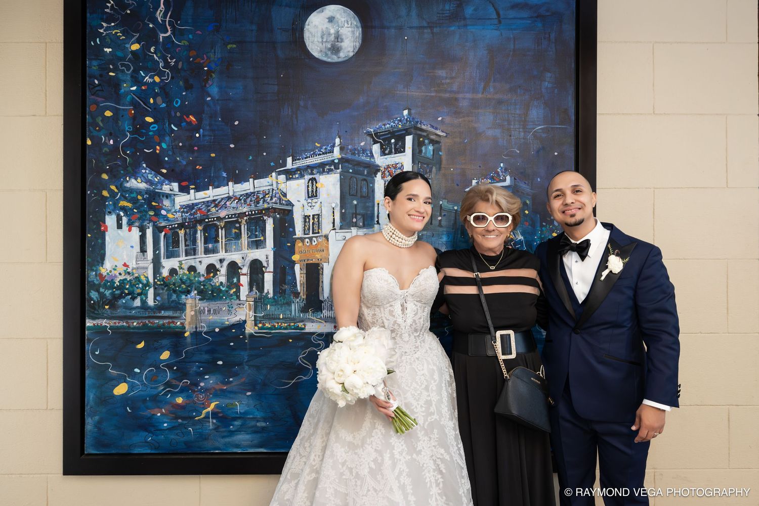 Bride and groom pose with a guest in front of a large moonlit painting at their elegant wedding reception.