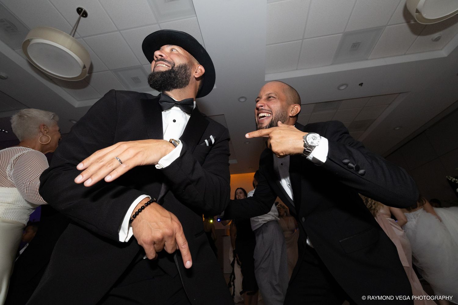 Two men in tuxedos laughing and dancing joyfully at a wedding reception party.