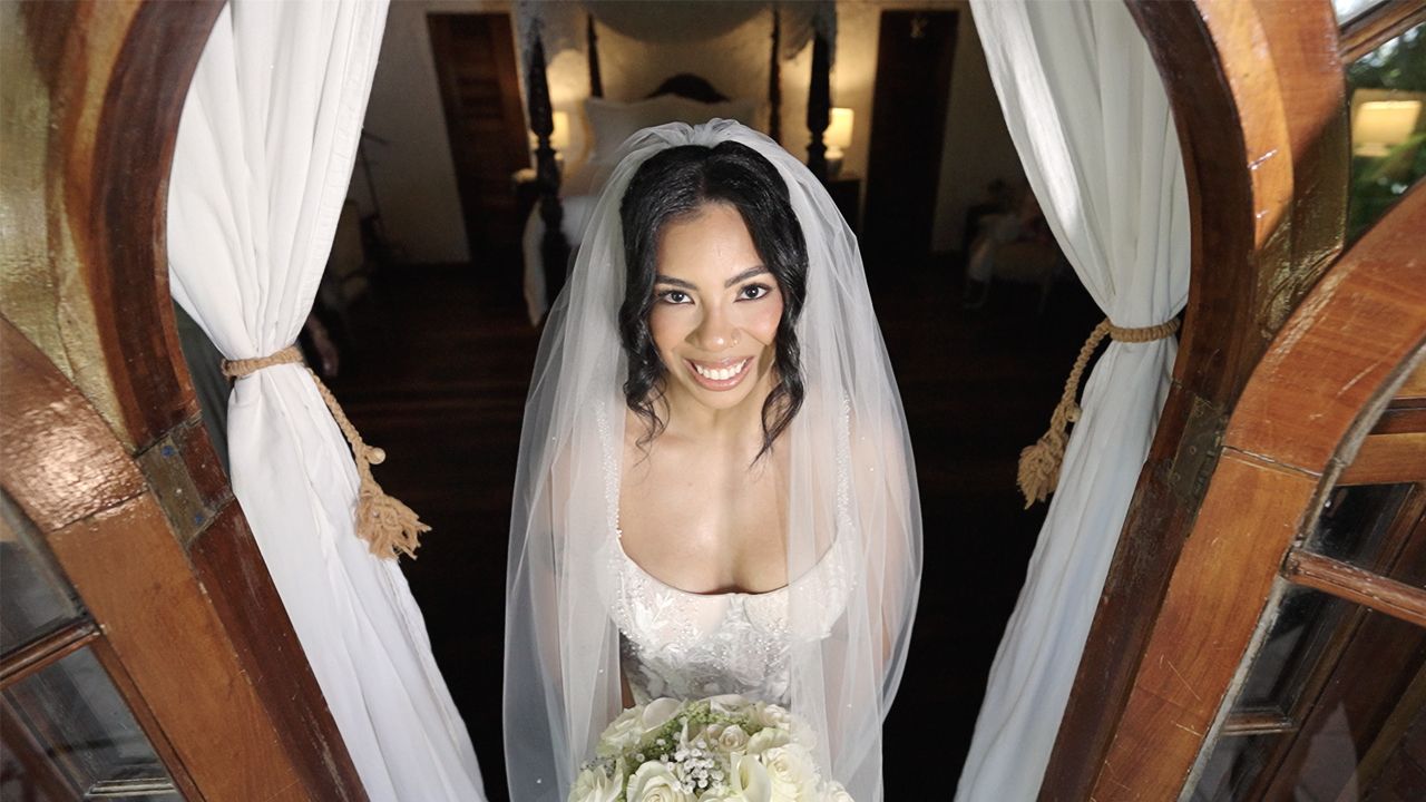 Radiant bride in white gown and veil smiles while holding a bouquet at the top of an elegant staircase.