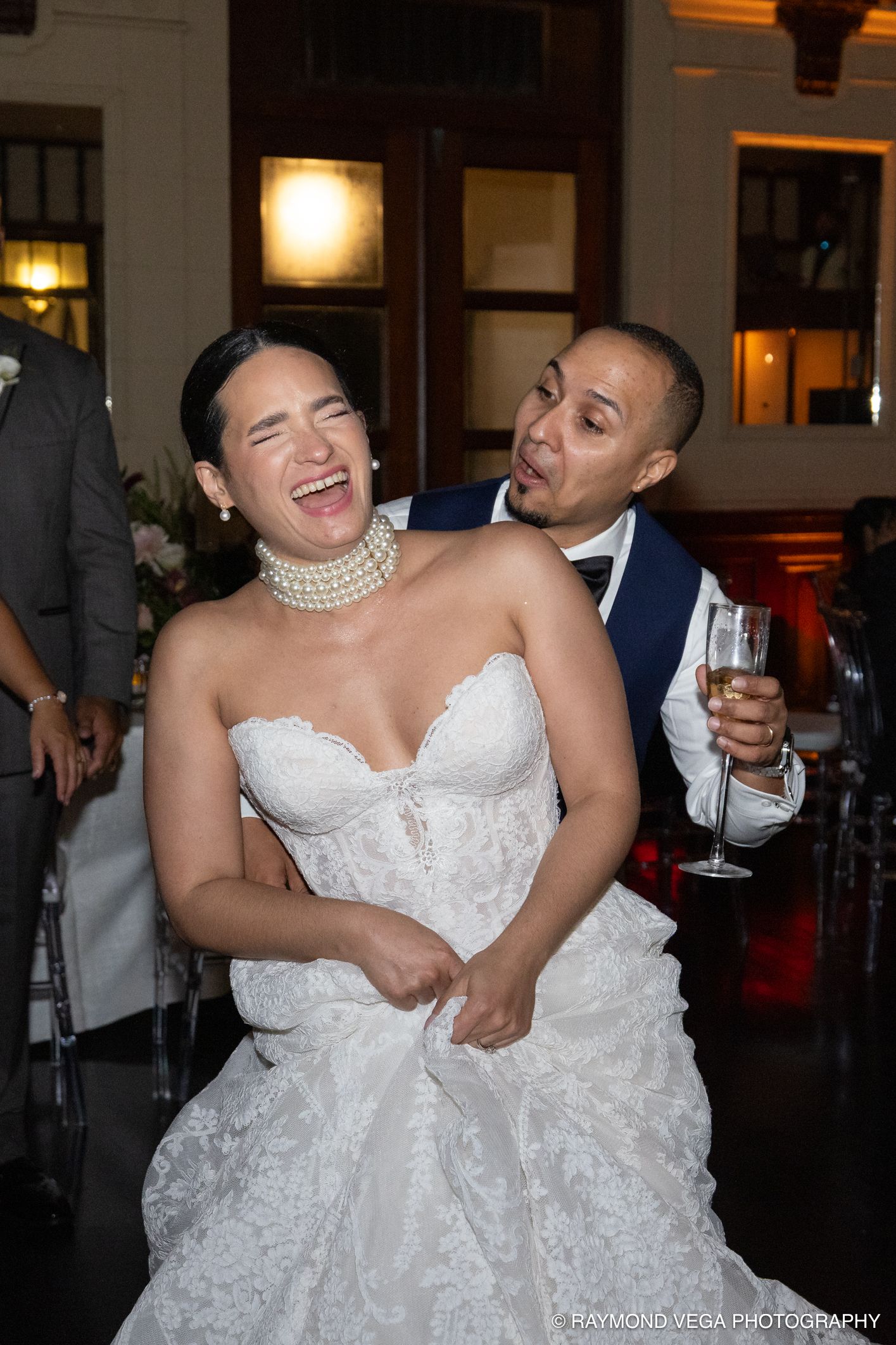 Joyful bride in white lace gown laughing with groom in navy suit holding champagne at wedding reception.