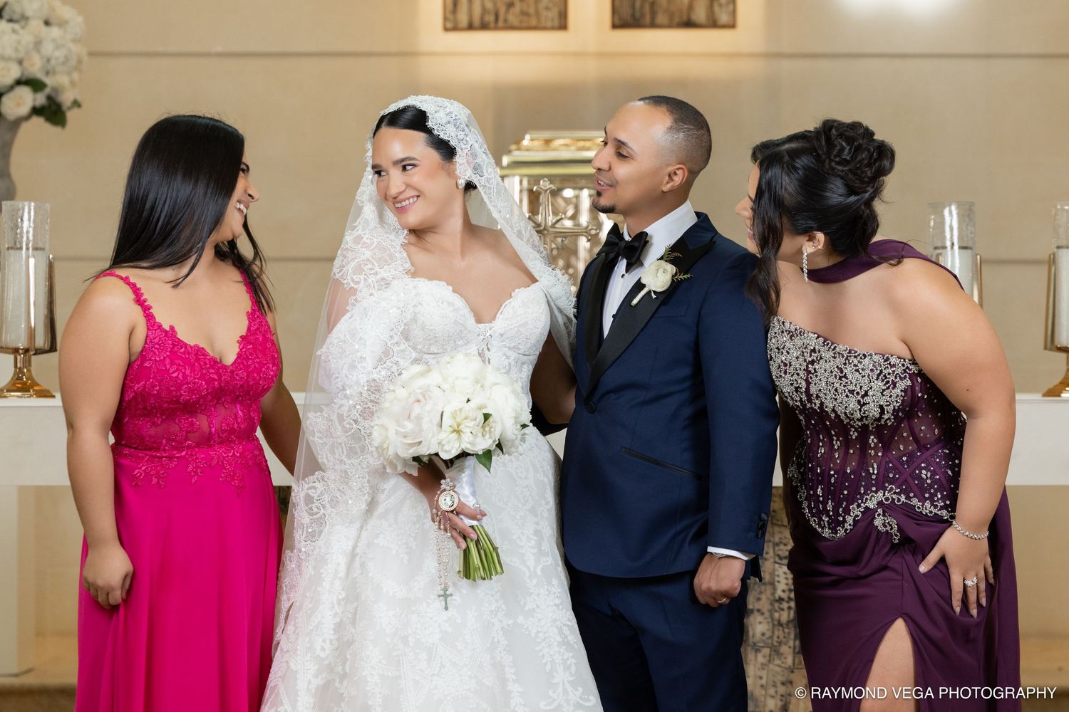 Bride in white lace gown and veil holds bouquet beside groom in navy suit, flanked by bridesmaids in pink and purple.