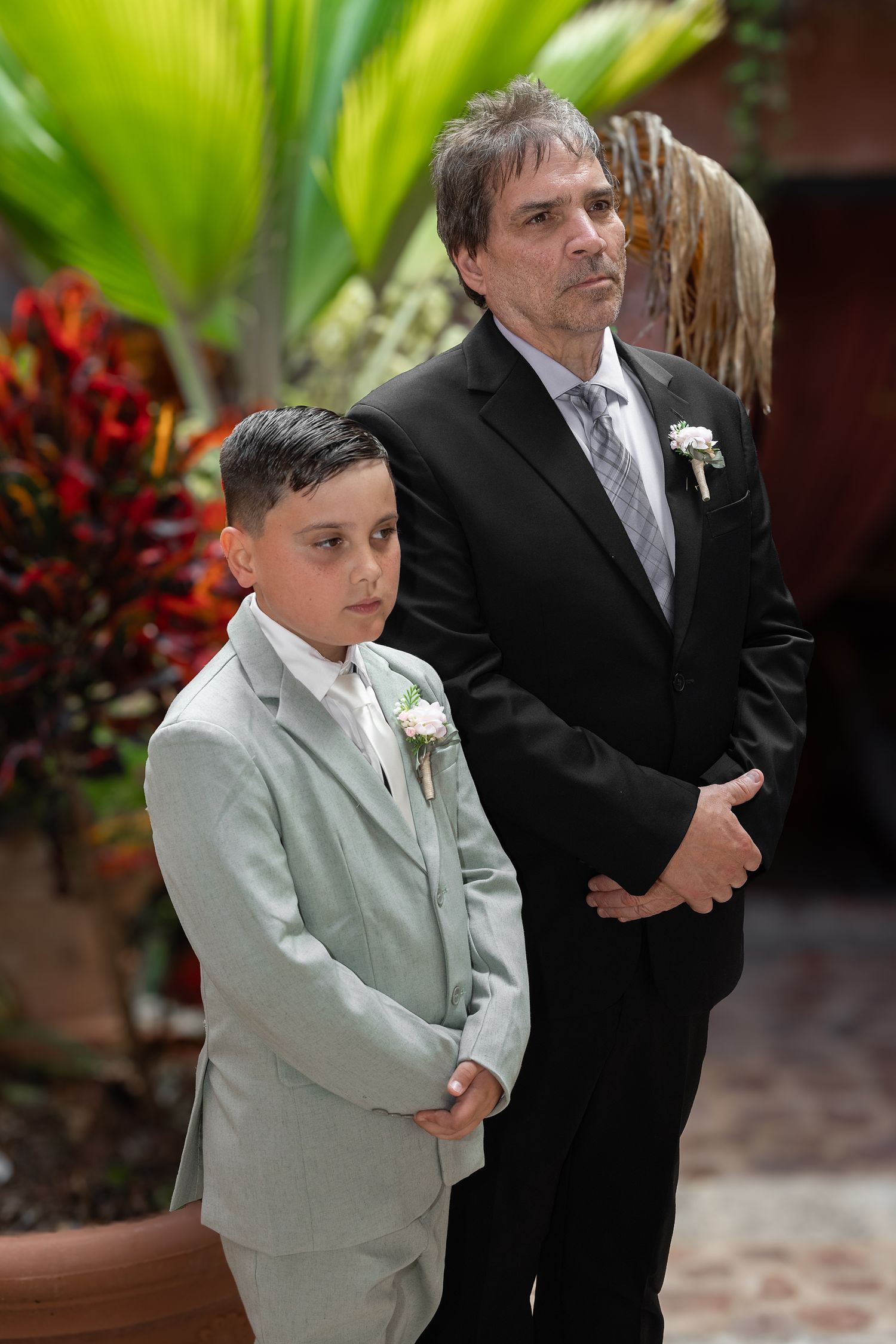 A boy and man in formal suits with boutonnieres stand attentively at a wedding ceremony outdoors.