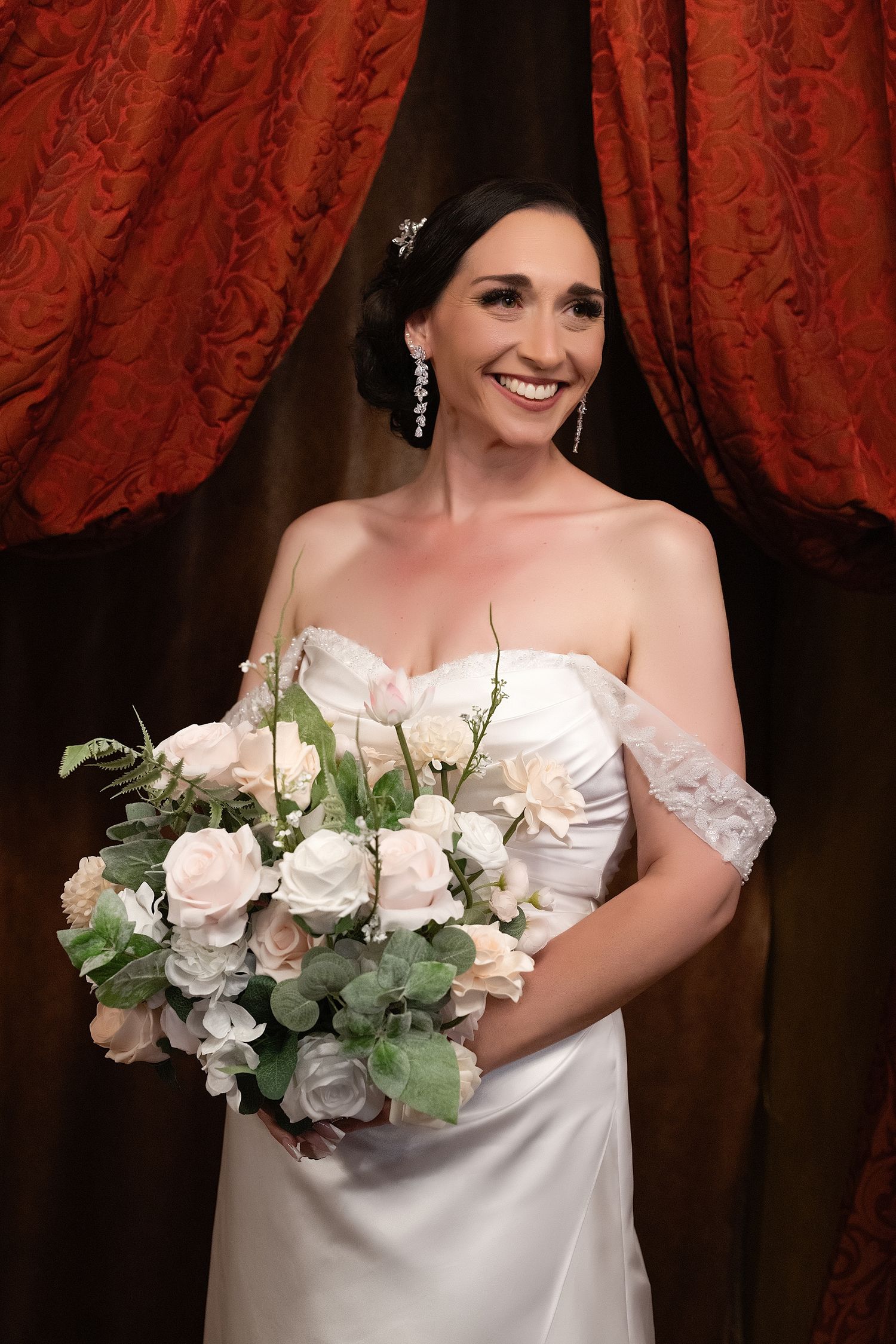 Smiling bride in white off-shoulder gown holds bouquet of white roses and greenery before red curtain backdrop.