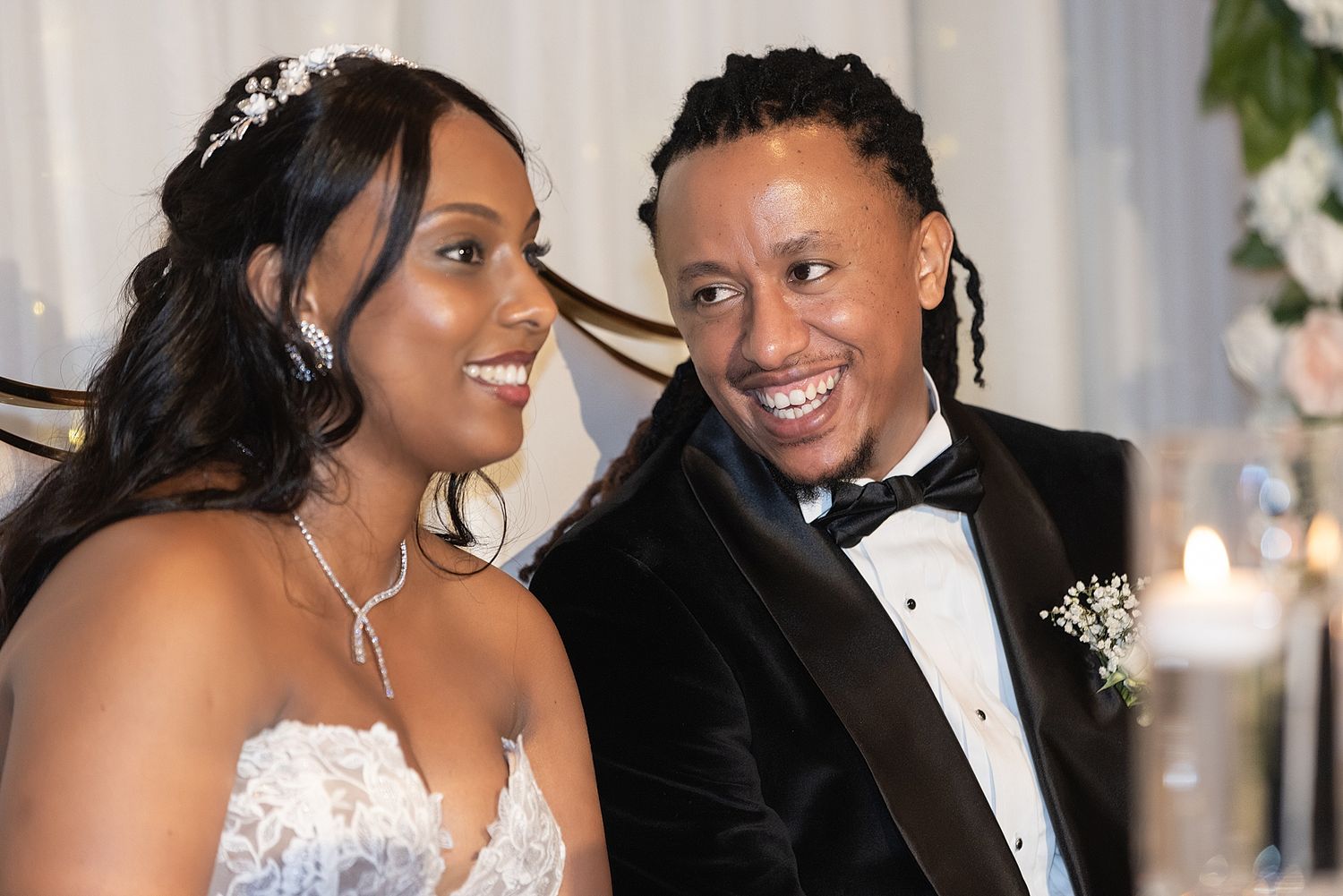A joyful couple in wedding attire smiles together at their reception, bride in lace gown and groom in black tuxedo.