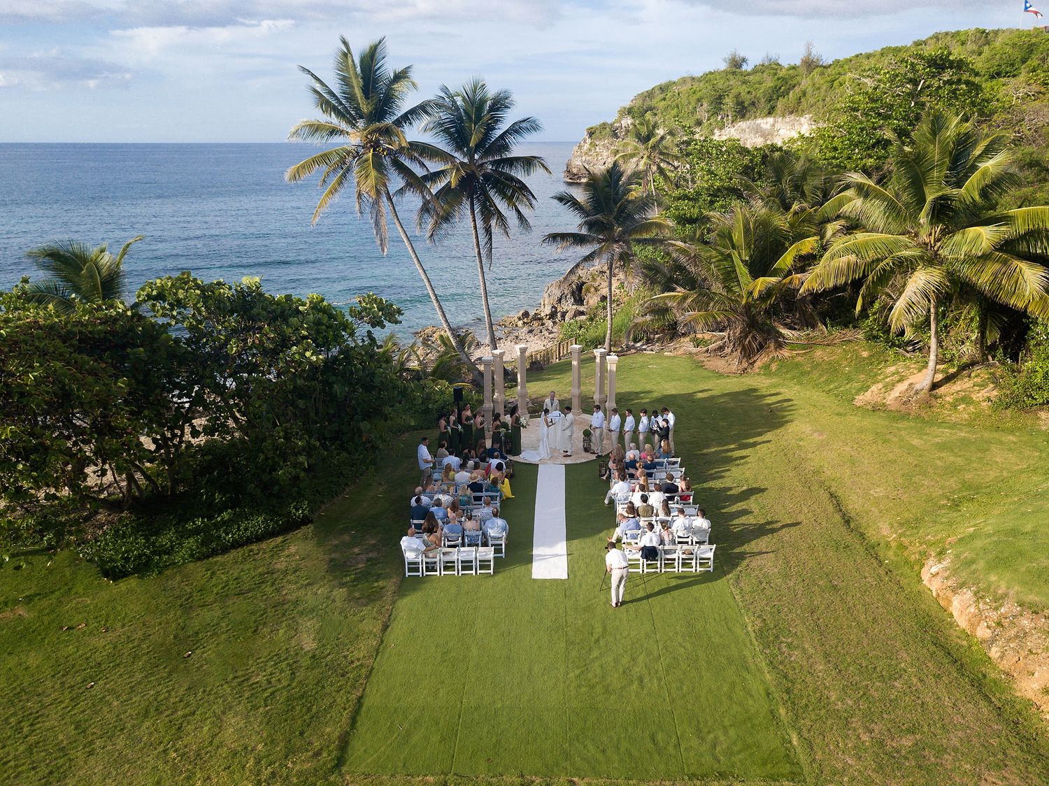 Aerial view of intimate oceanfront wedding ceremony on tropical lawn with palm trees and blue sea.