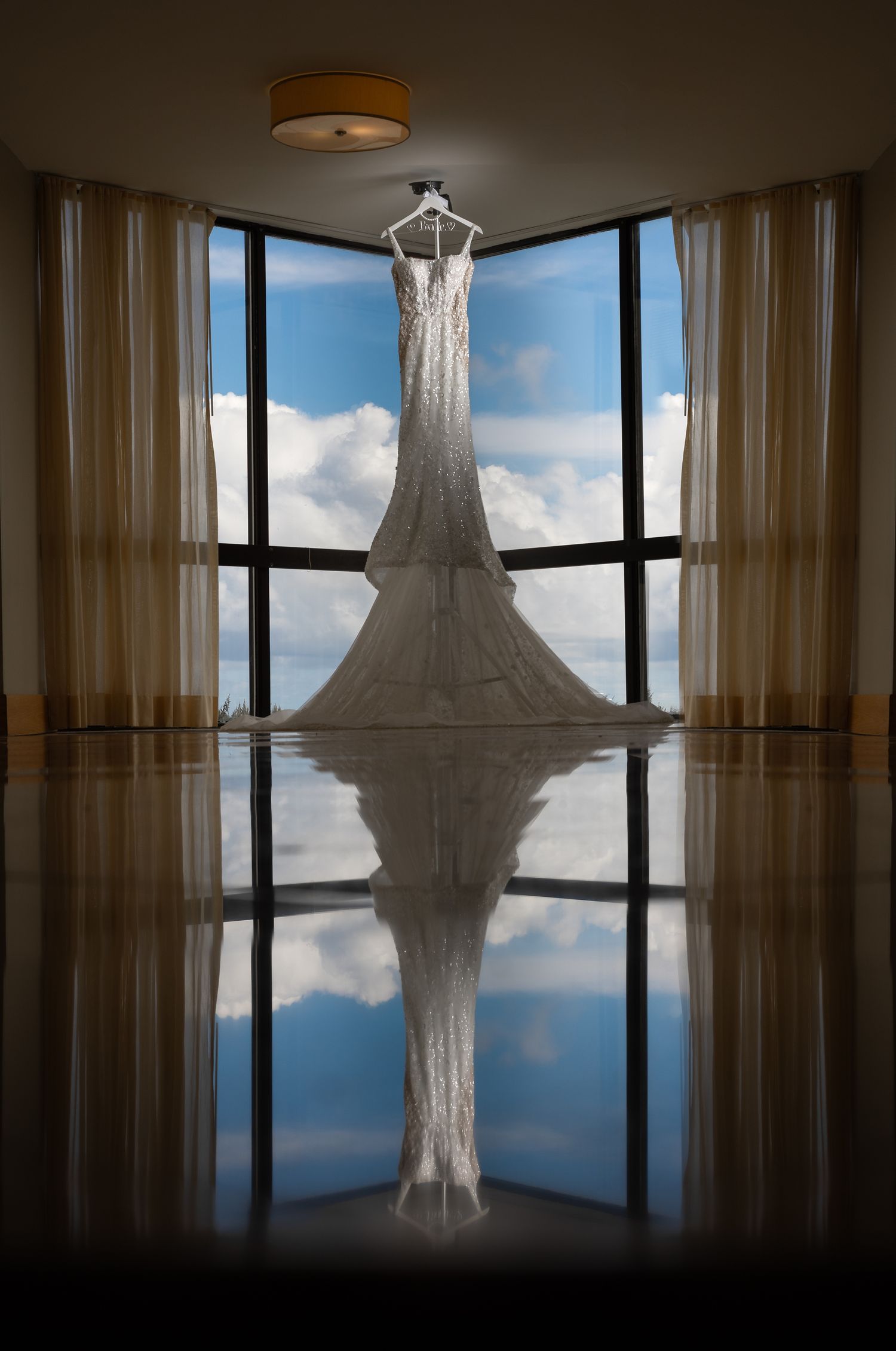 Elegant wedding dress hanging in a sunlit corner window, reflected on polished floor with blue sky backdrop.