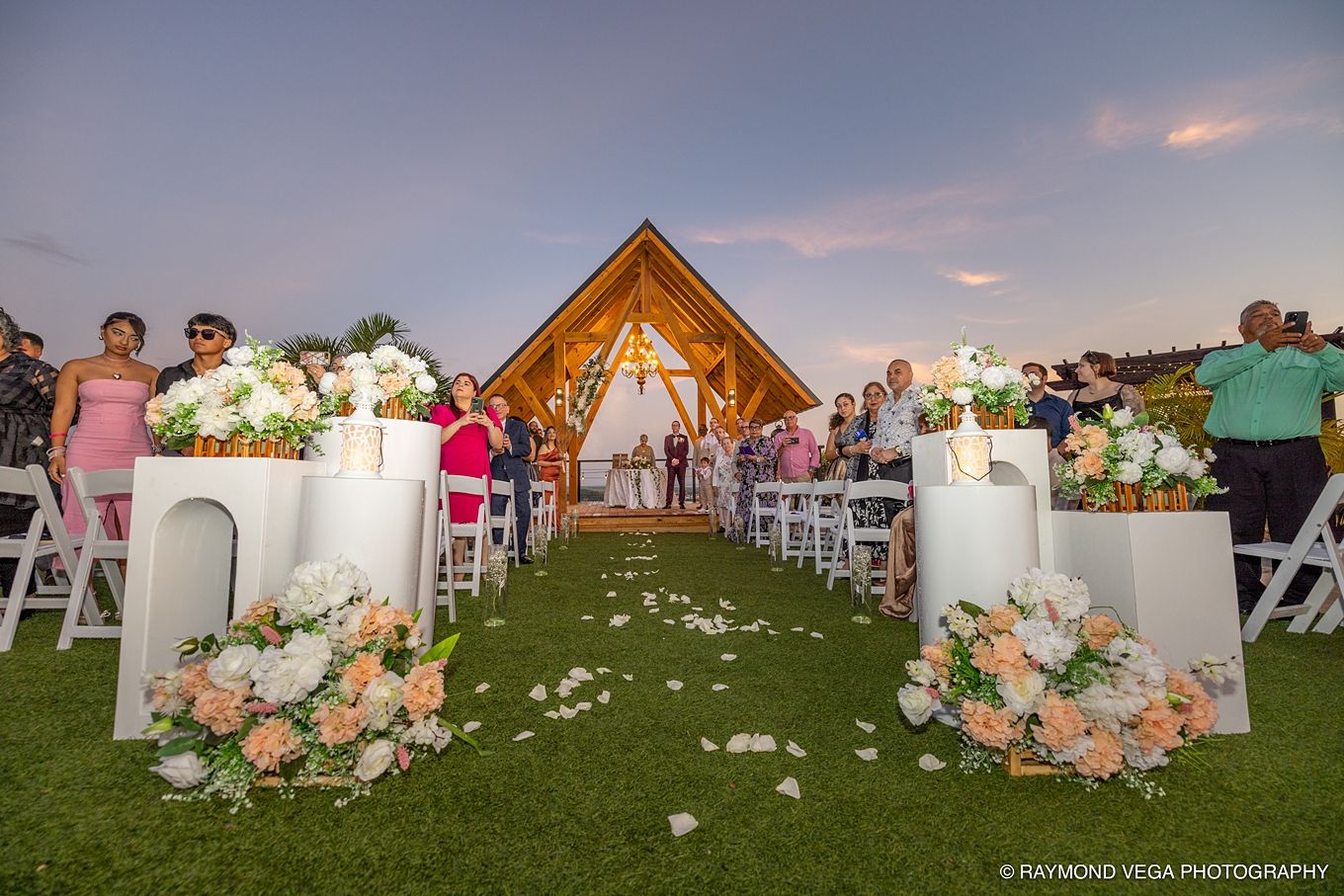 Outdoor wedding ceremony at sunset with floral arrangements lining the aisle leading to a wooden arch chapel.