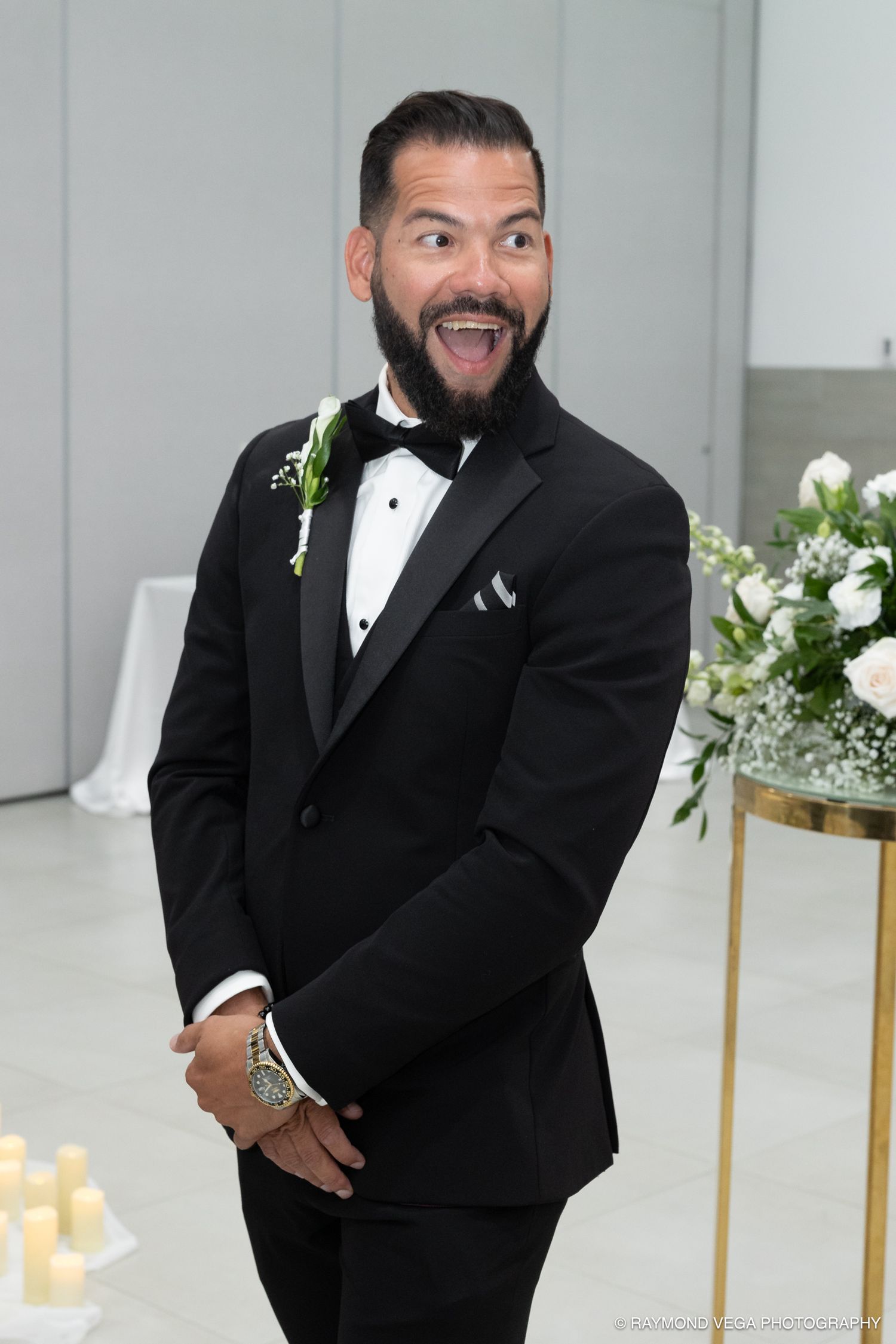 Groom in black tuxedo with bow tie and boutonniere smiling joyfully at wedding ceremony altar.
