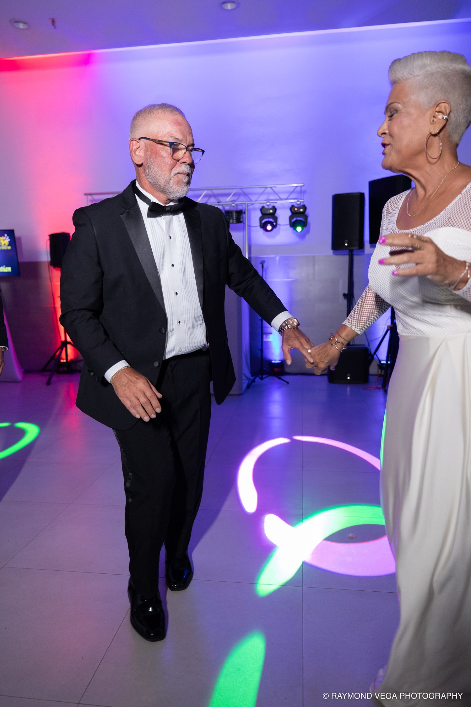 Elderly couple dancing elegantly at a formal event, man in tuxedo and woman in white gown, colorful dance floor lights.
