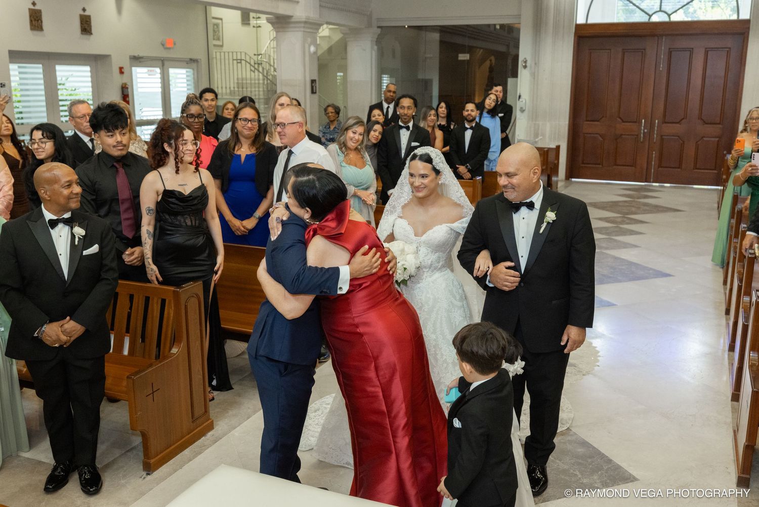Bride walks down church aisle hugging guest in red dress, surrounded by wedding party and guests in formal attire.