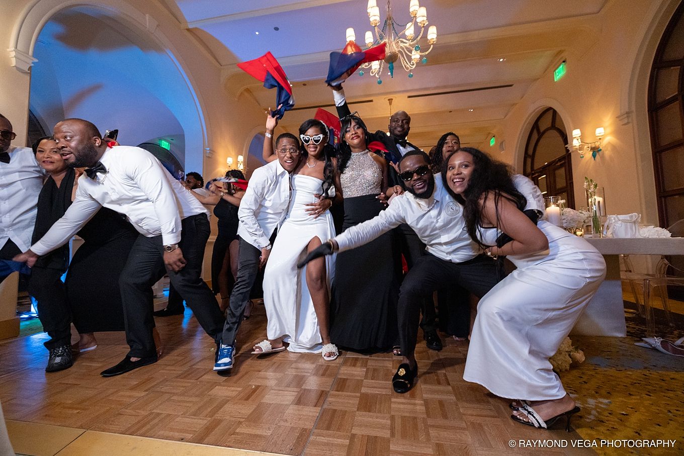 Wedding party dancing joyfully in elegant ballroom, guests in white and black attire celebrating with flags on dance floor.