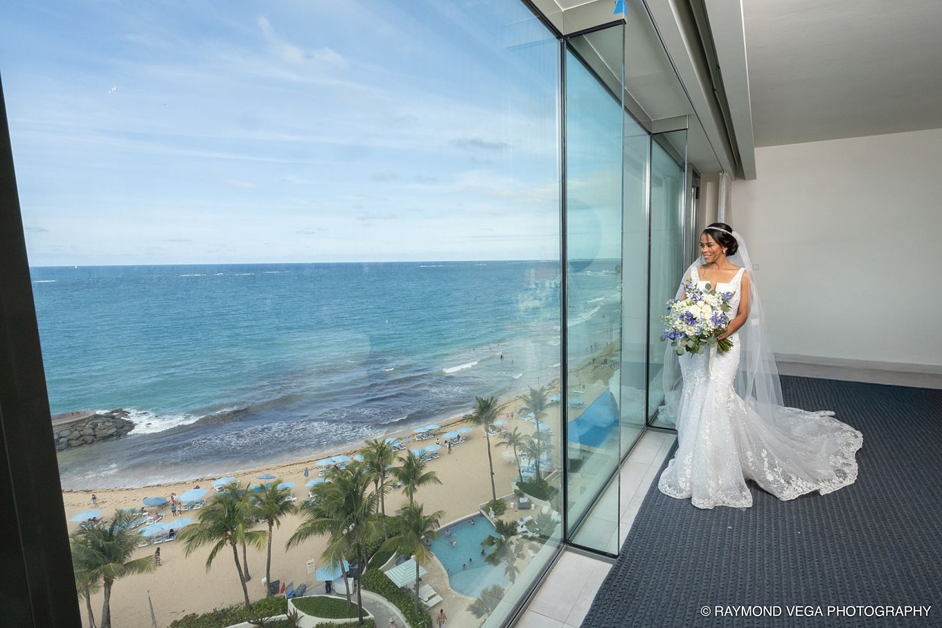 Bride in white wedding gown poses by floor-to-ceiling glass windows overlooking a tropical beach and ocean view. Hotel la Concha Wedding