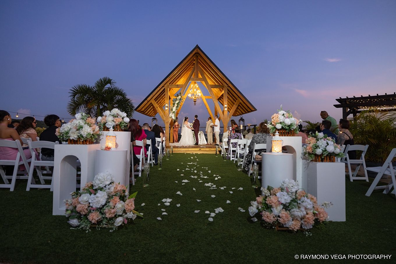 Elegant outdoor wedding ceremony at dusk with floral arrangements, candles, and a lit pavilion altar surrounded by seated guests.