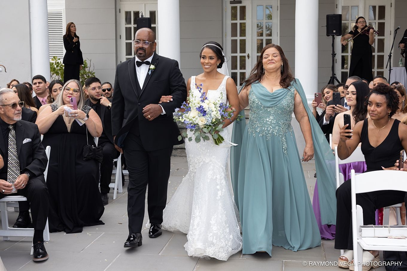 Bride in white gown walks down aisle with parents at outdoor wedding ceremony, guests watching and smiling.