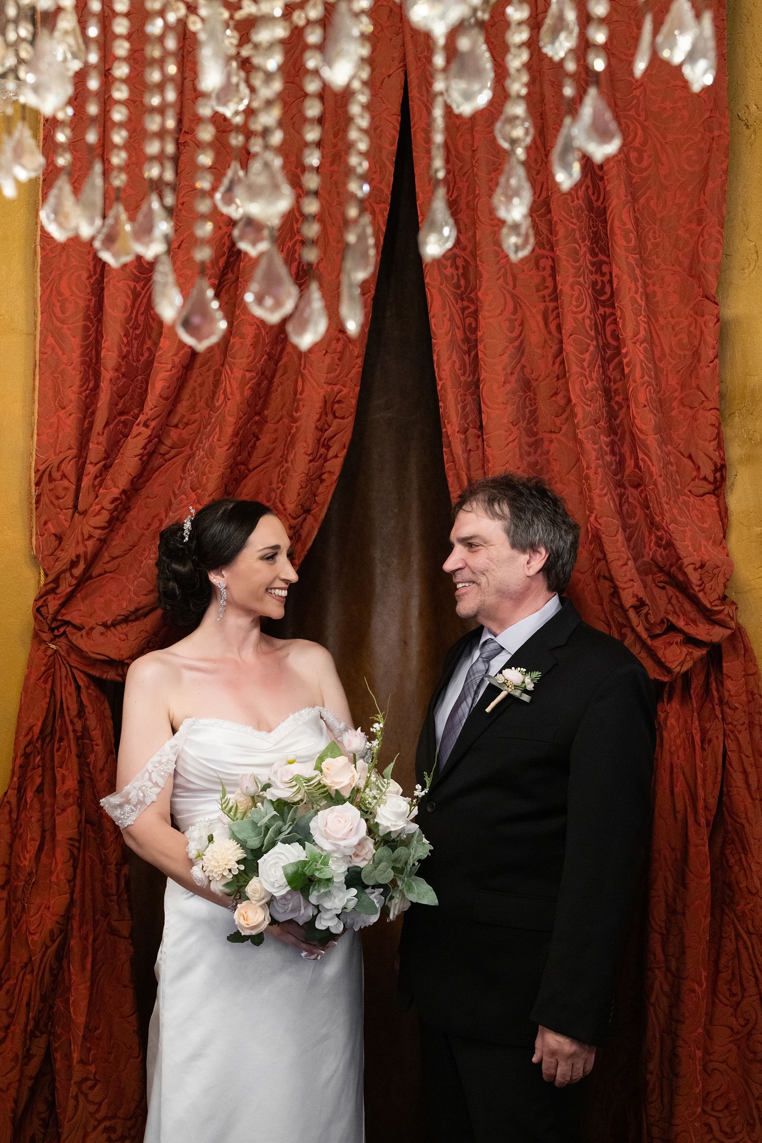 Bride and groom smiling at each other under crystal chandelier, framed by elegant red velvet curtains, bride holding white roses.