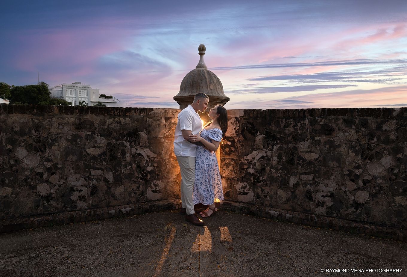 Couple embracing at sunset on historic stone fortress wall in Old San Juan, Puerto Rico, with colorful pink and purple sky - Love Story