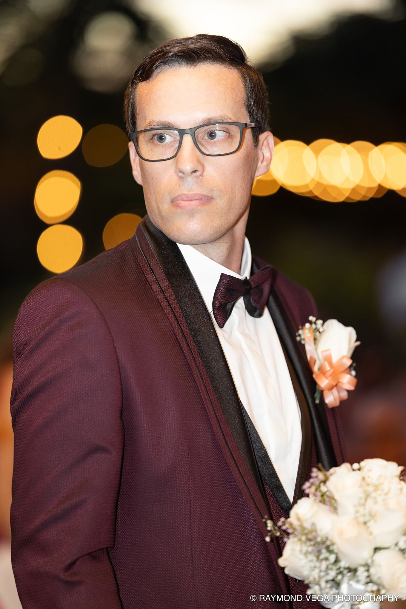Groom in burgundy tuxedo with black lapels, bow tie, boutonniere, and glasses holding bouquet at wedding reception.