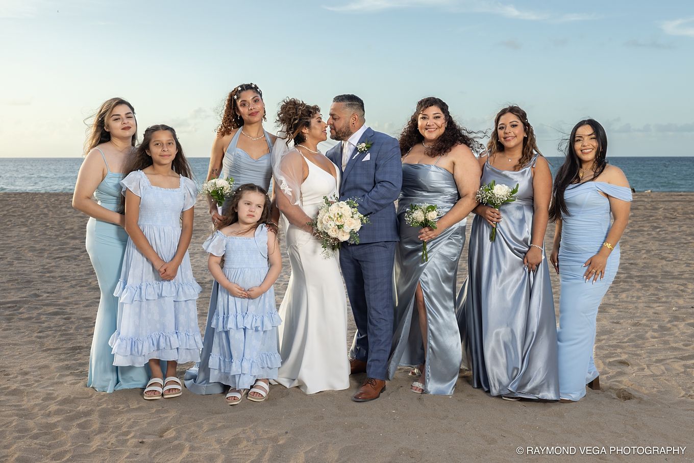 Beach wedding party in light blue dresses with bride and groom kissing, ocean backdrop at sunset.
