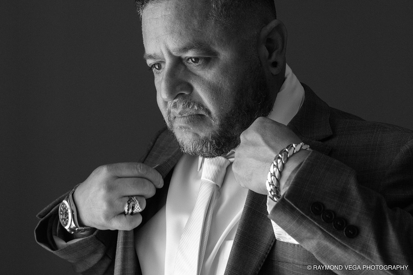 Black and white portrait of a man in a suit adjusting his tie, wearing a watch and ring, studio photography.