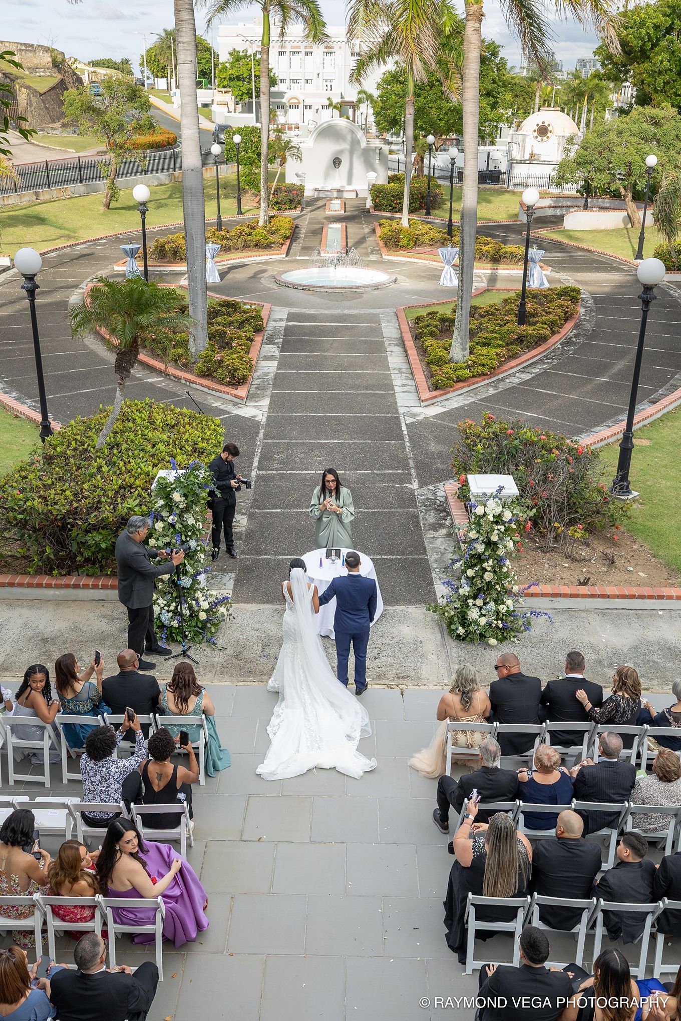 Aerial view of an outdoor wedding ceremony with bride and groom walking the aisle, guests seated, and a fountain backdrop. Antiguo Casino