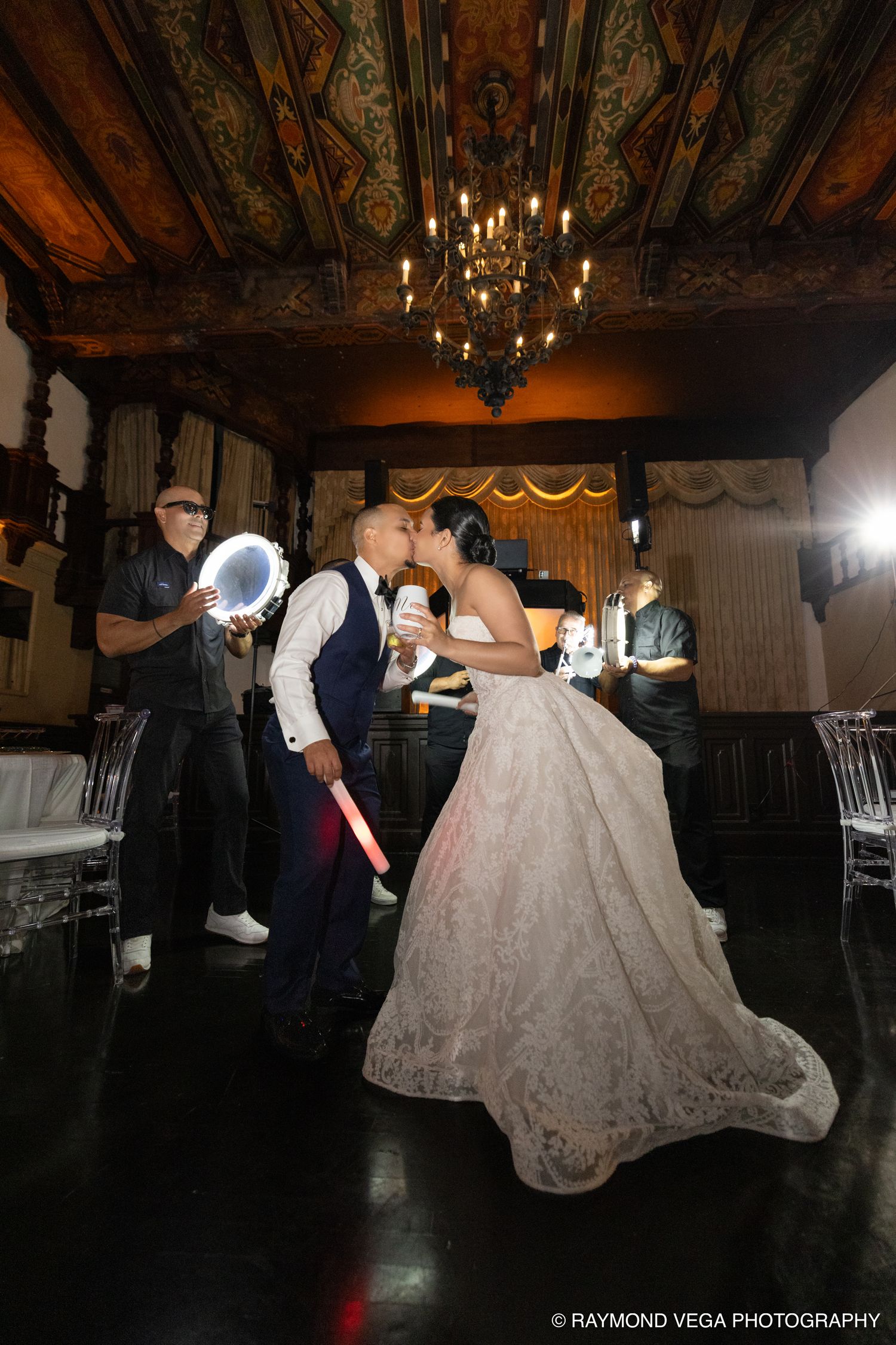 Bride and groom share a kiss on the dance floor during their elegant wedding reception in an ornate ballroom.
