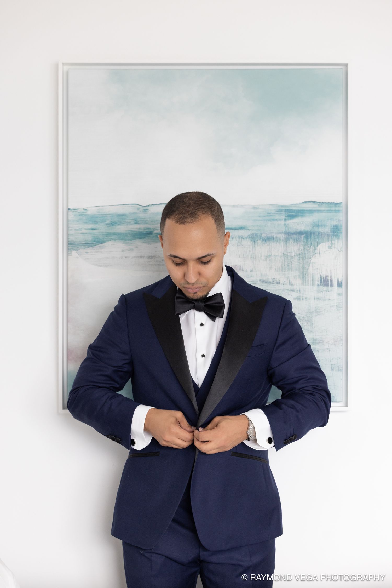 Groom in navy blue tuxedo with black bow tie buttoning jacket before wedding ceremony.