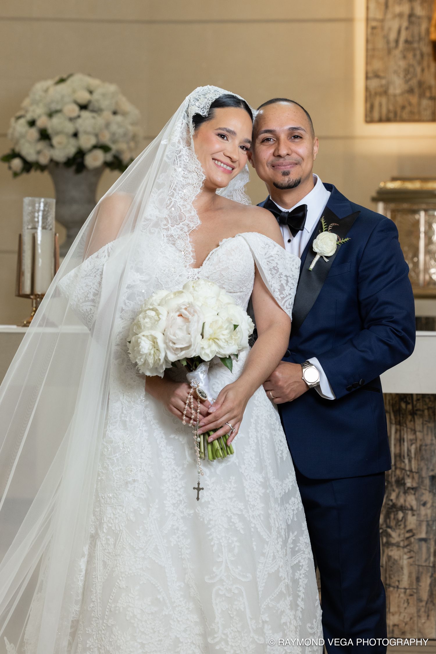Bride in lace gown with veil holds white bouquet beside groom in navy suit at elegant church wedding ceremony.