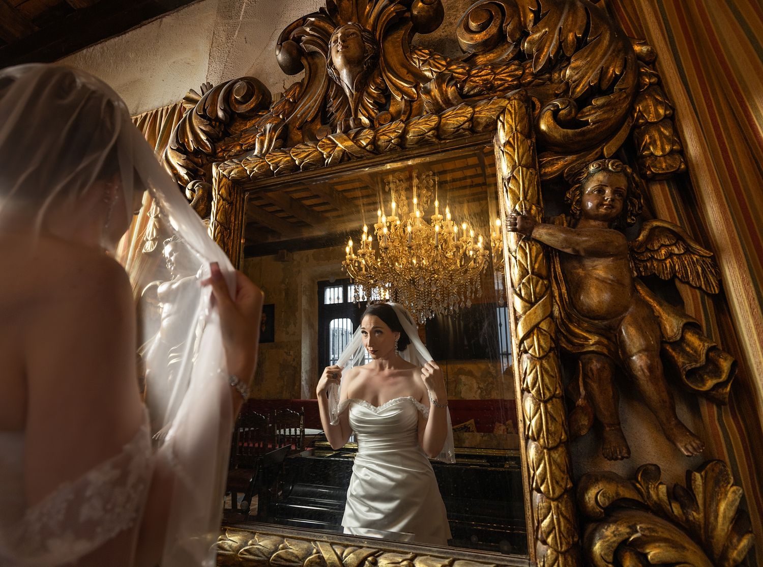 Bride in white gown adjusts veil before ornate gold baroque mirror with cherub sculpture in elegant venue.