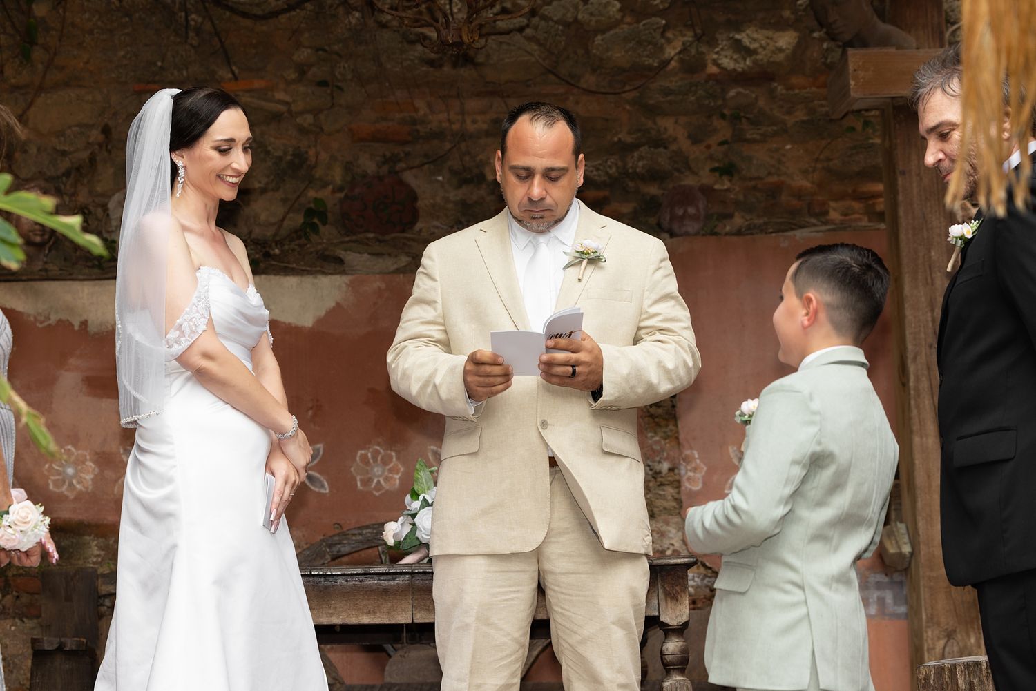 Groom in beige suit reads wedding vows to bride in white dress and veil during outdoor rustic ceremony. The Cannon Club in Old San Juan