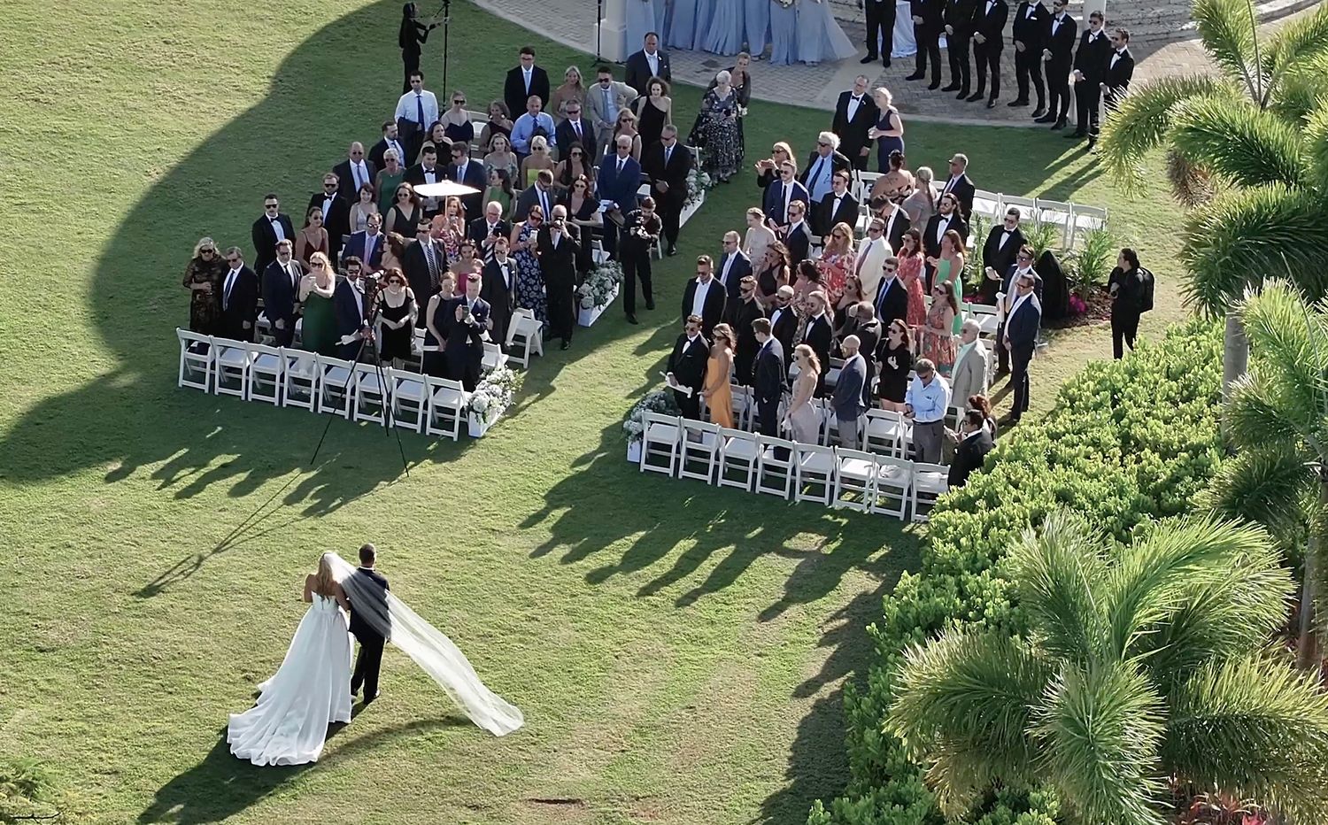 Aerial view of outdoor wedding ceremony on golf course lawn with white chairs and tropical palms.