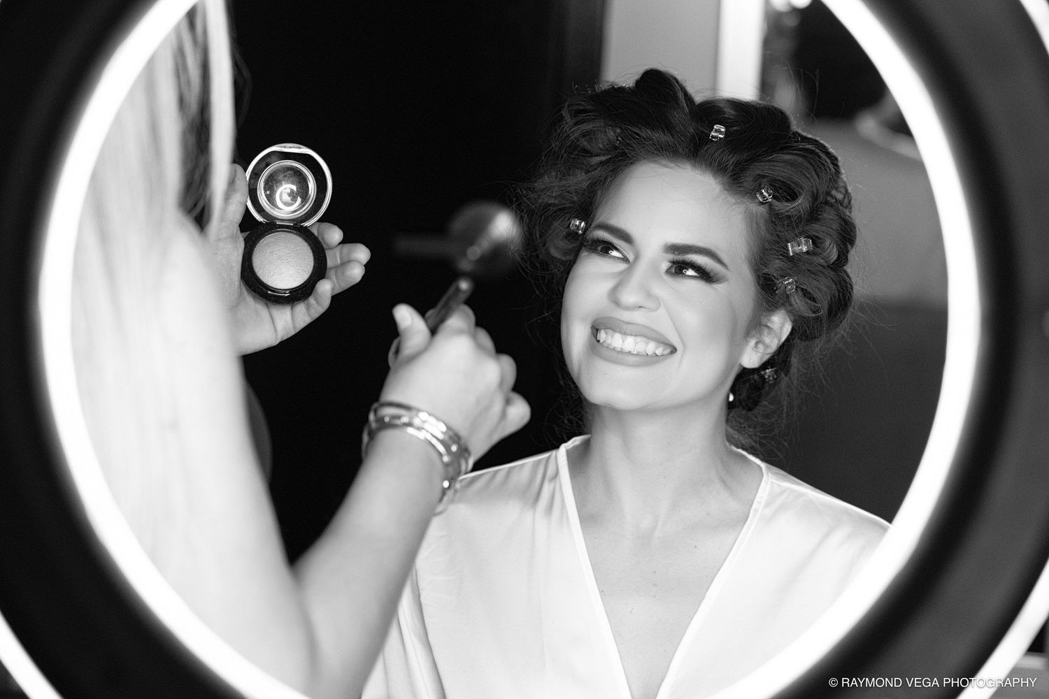 Black and white photo of a smiling bride applying makeup with a powder brush in front of a round lighted mirror.