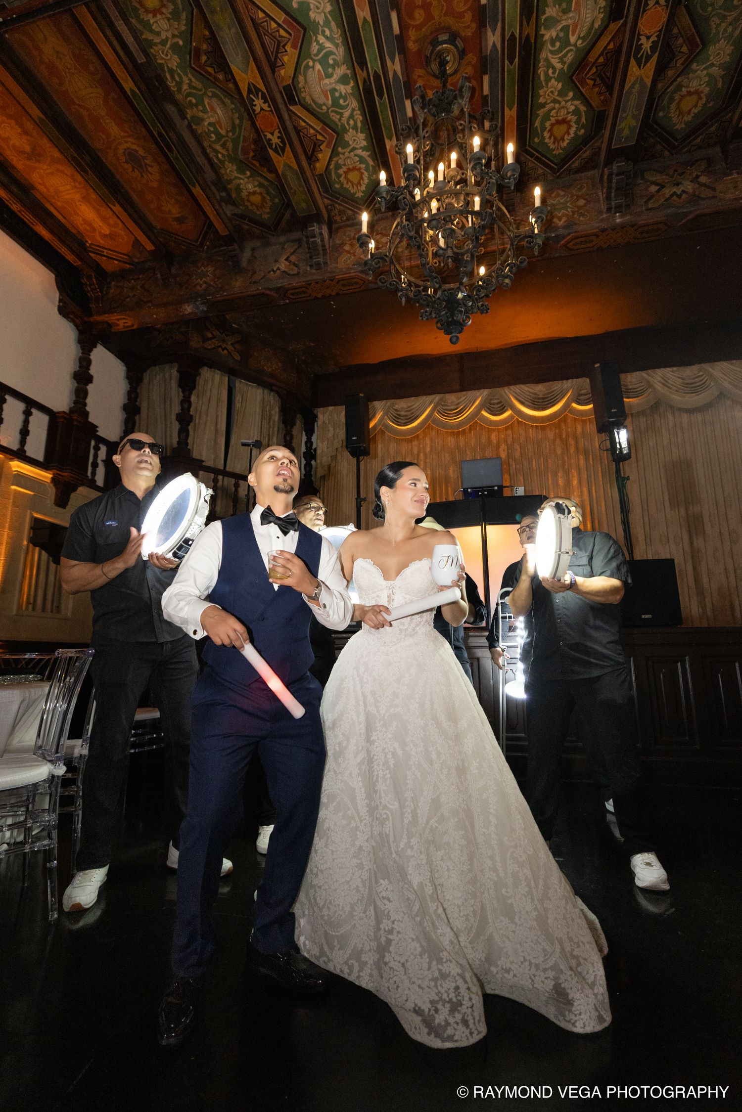 Bride and groom dancing joyfully at elegant wedding reception, holding light wands under ornate chandelier.
