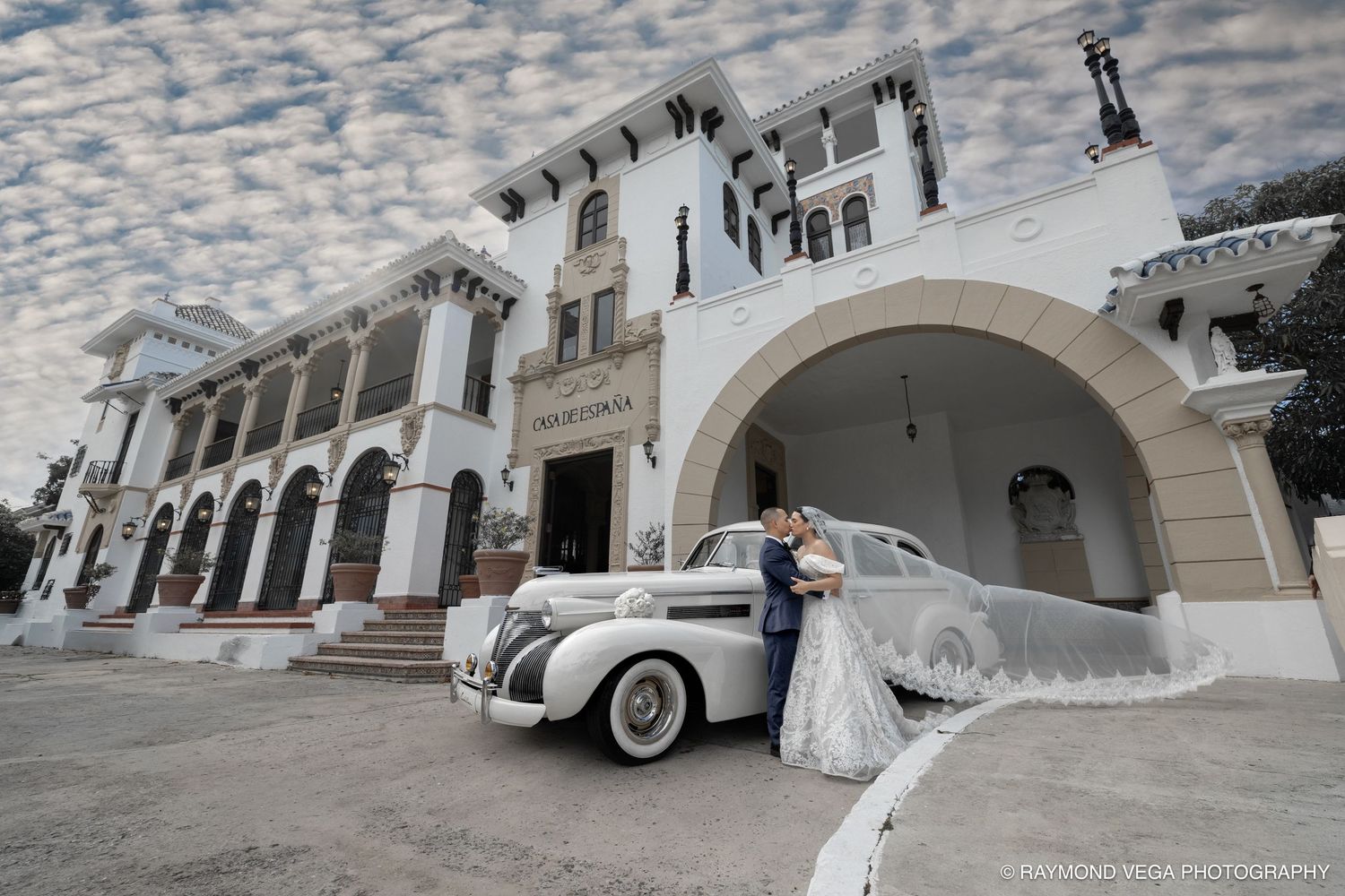 Bride and groom pose beside a vintage white car in front of an elegant white colonial building under a dramatic cloudy sky.