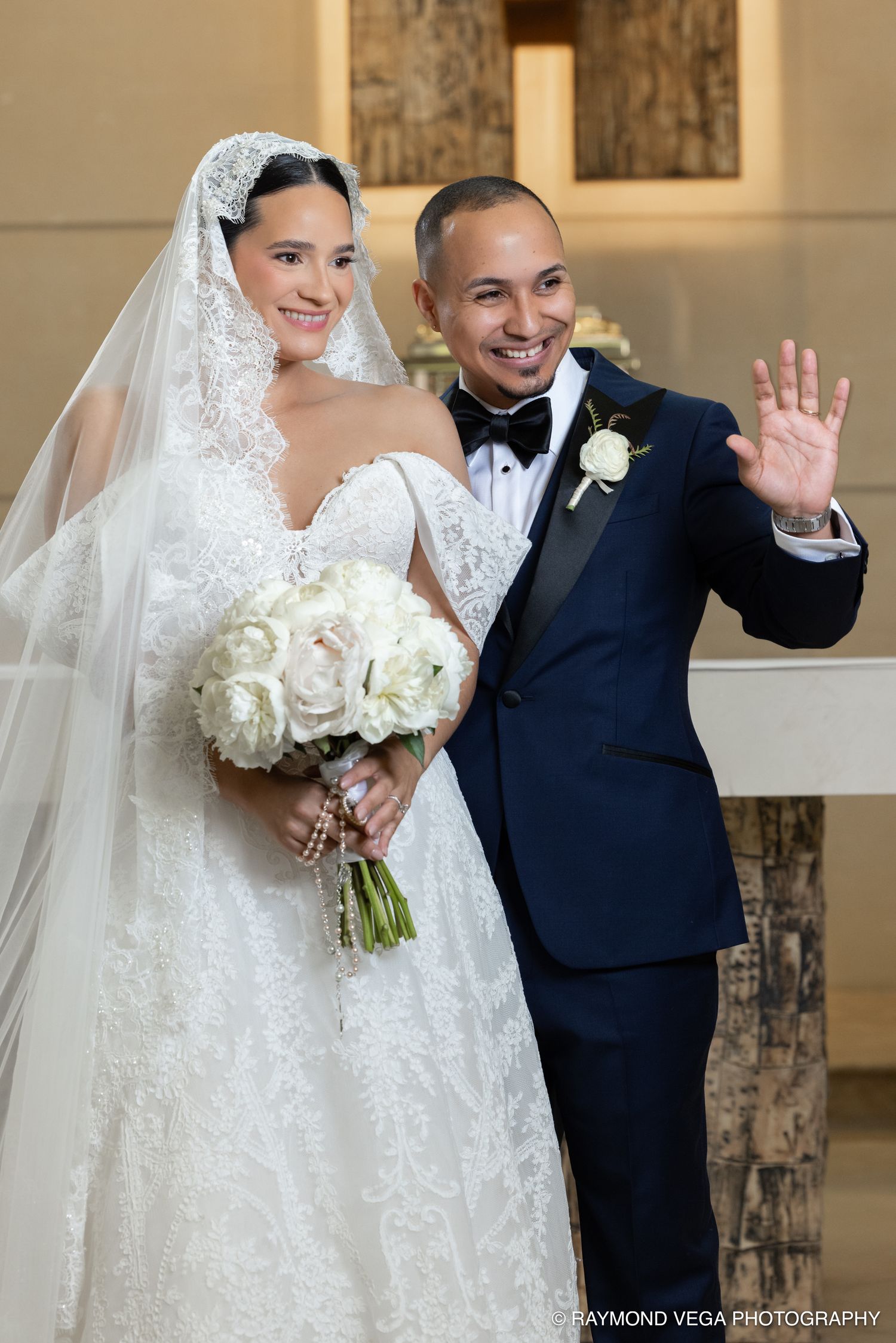 Bride in lace gown with veil holds white bouquet beside groom in navy tuxedo waving at wedding ceremony.