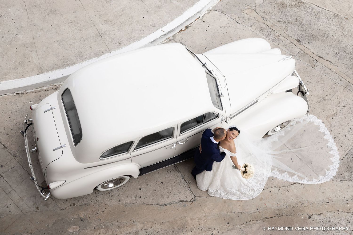 Aerial view of bride and groom posing beside a classic white vintage car on their wedding day.