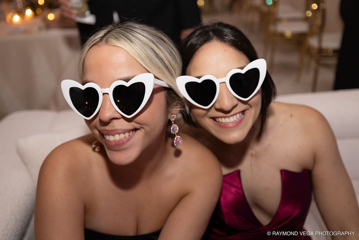 Two women smiling in white heart-shaped sunglasses at a formal event, wearing elegant dresses.