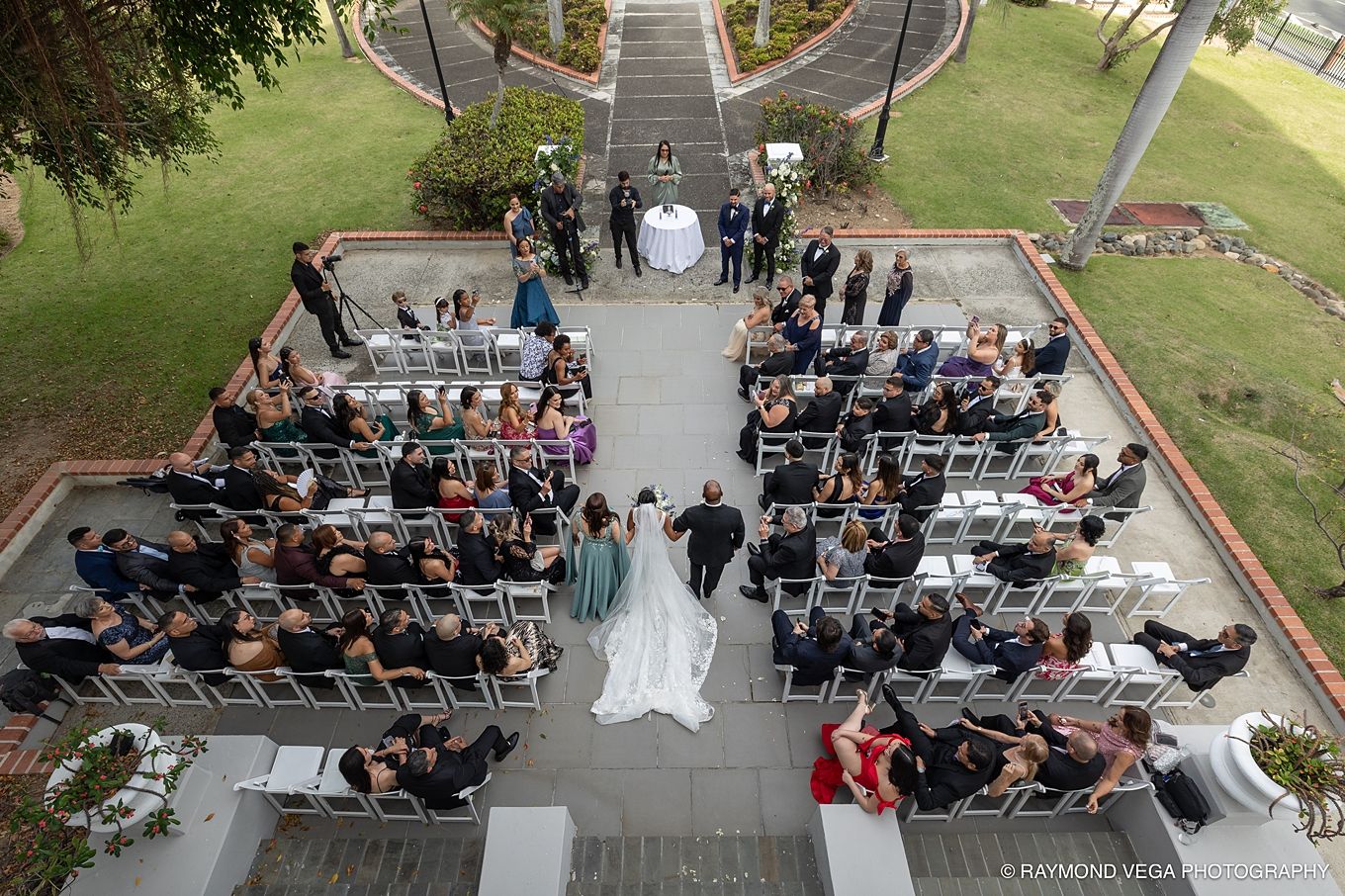 Aerial view of an outdoor wedding ceremony with guests seated in white chairs on a patio surrounded by greenery. Antiguo Casino Puerto Rico
