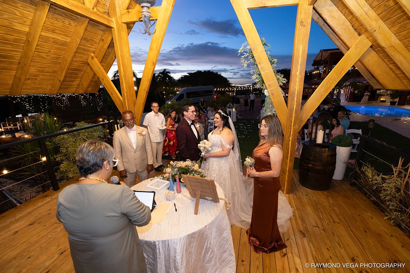 Outdoor wedding ceremony on a wooden deck at dusk with bride, groom, and officiant under a timber pergola.