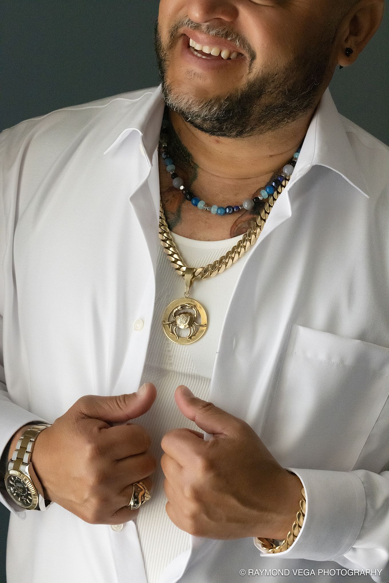 Smiling man in white shirt wearing gold chain necklace, beaded necklaces, gold bracelet, and watch, adjusting his shirt.