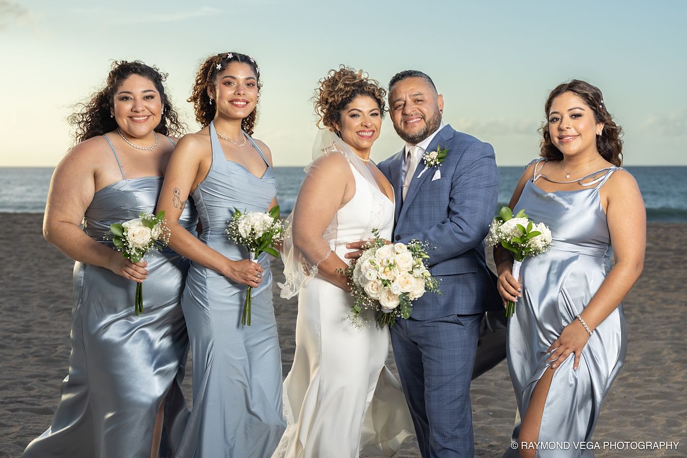 Bride and groom pose with three bridesmaids in blue satin dresses on a beach at sunset, holding white bouquets at Courtyard by Marriott