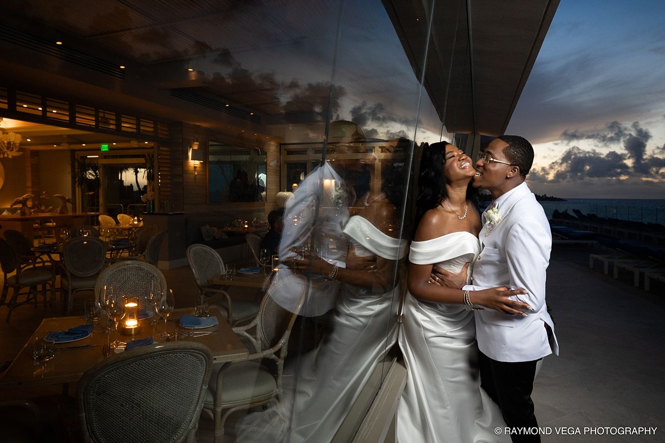 Bride and groom share a romantic kiss reflected in glass at a sunset oceanview venue.