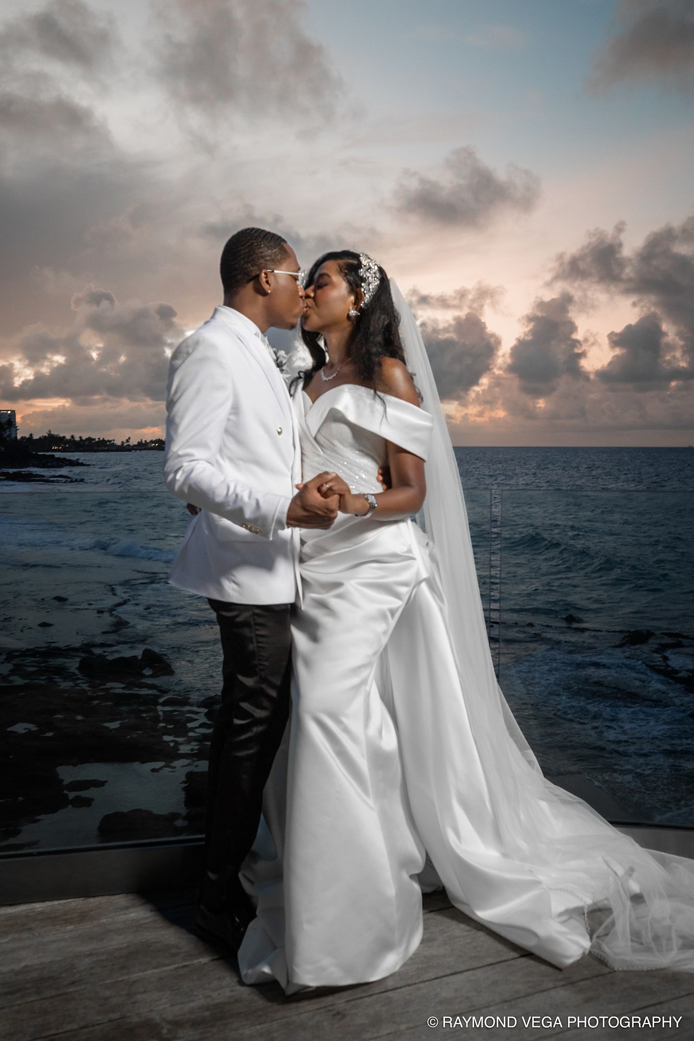 Bride and groom share a romantic kiss at sunset by the ocean, bride in elegant white gown with veil, groom in white tuxedo.