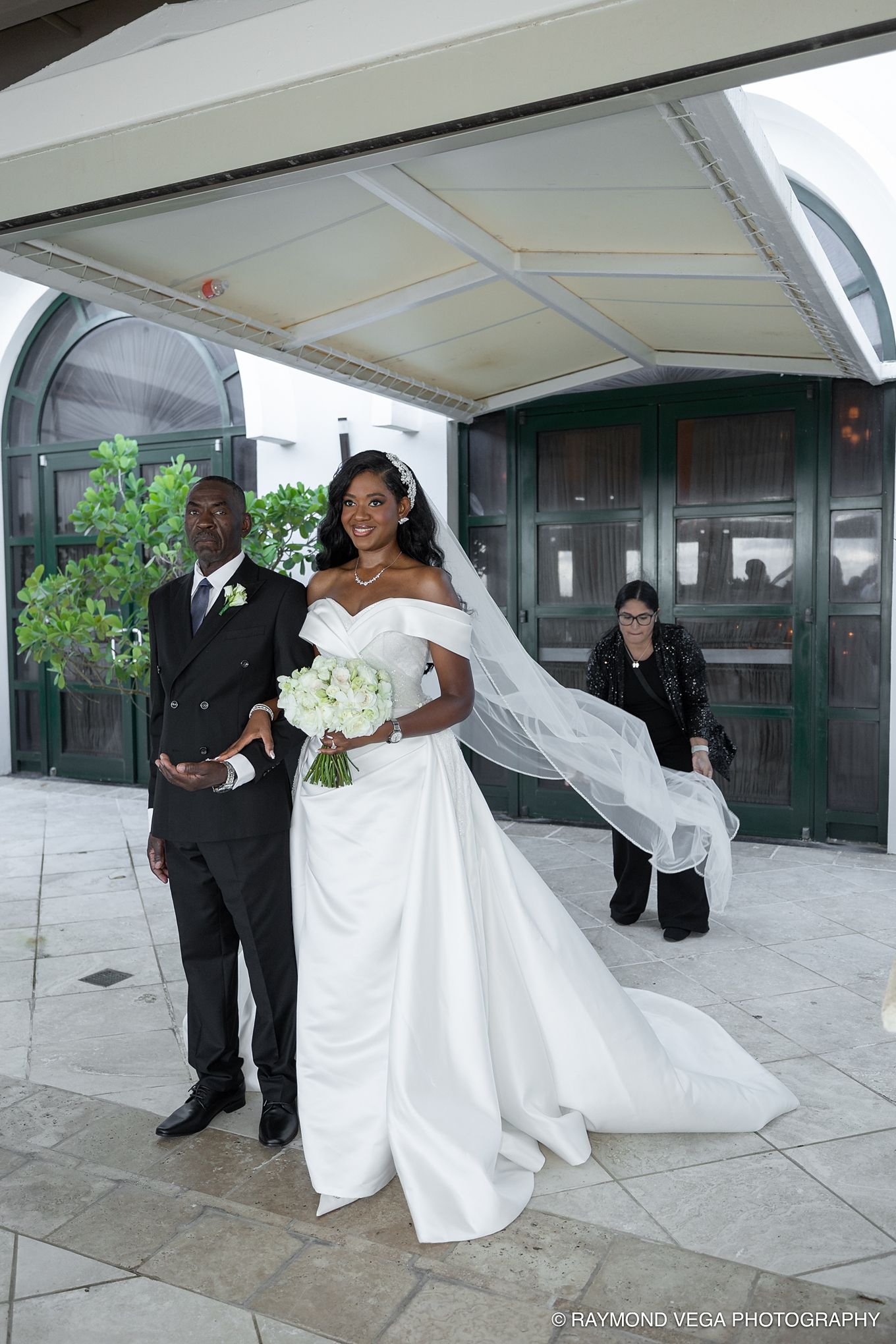 Bride in white off-shoulder gown with veil and bouquet stands with groom in black suit outside a church.