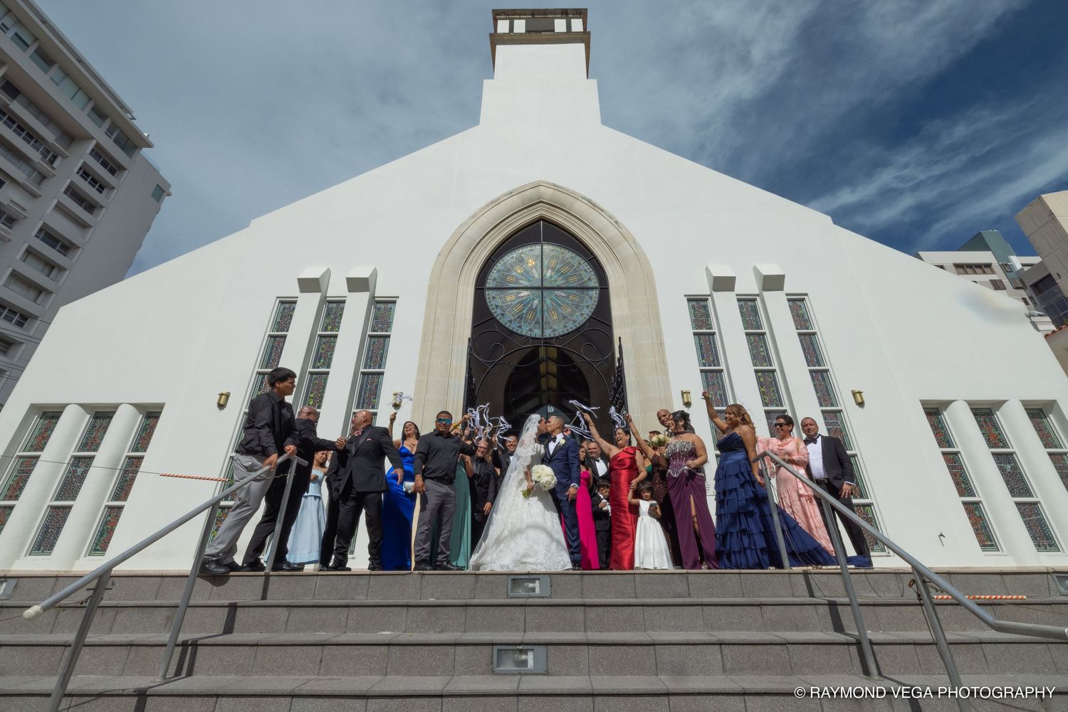 Wedding party posing on steps of a white church with gothic arch doorway under blue skies.