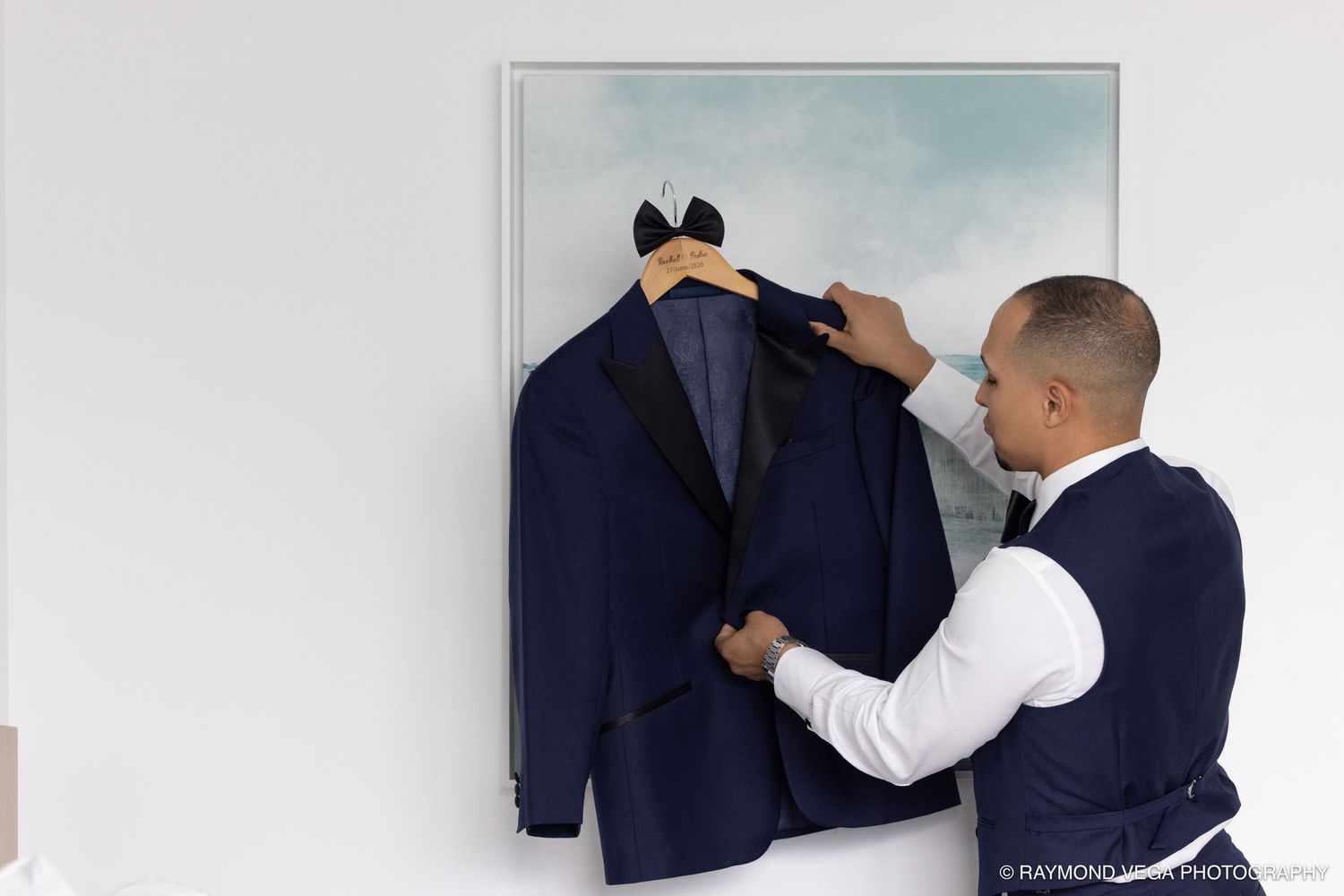 A groom in a vest hangs a navy blue tuxedo jacket on a hanger against a framed painting on a white wall.
