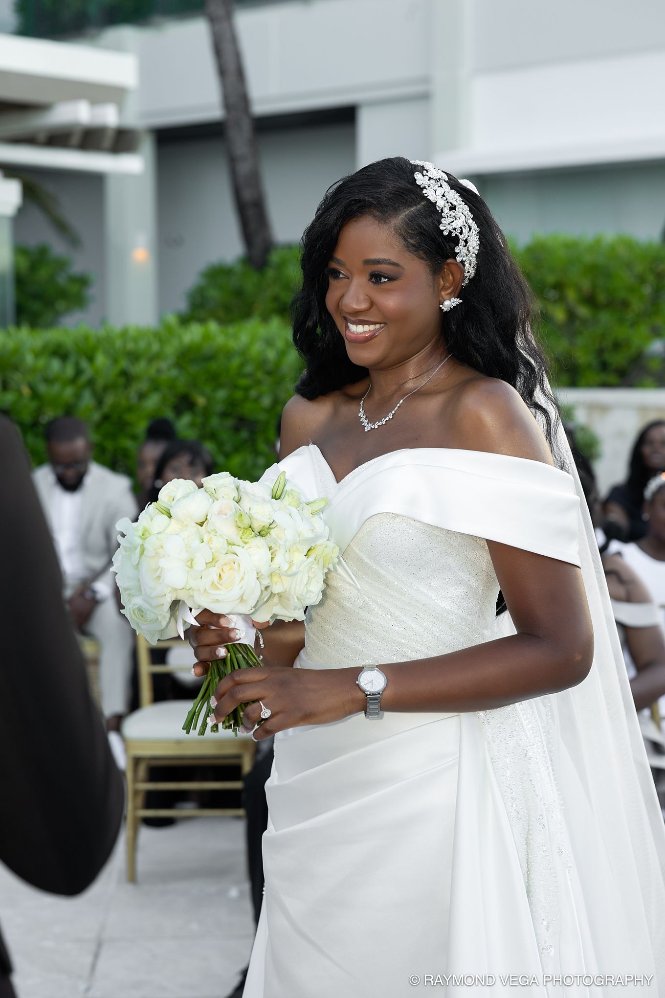 Smiling Black bride in off-shoulder white gown holds white bouquet at outdoor wedding ceremony.