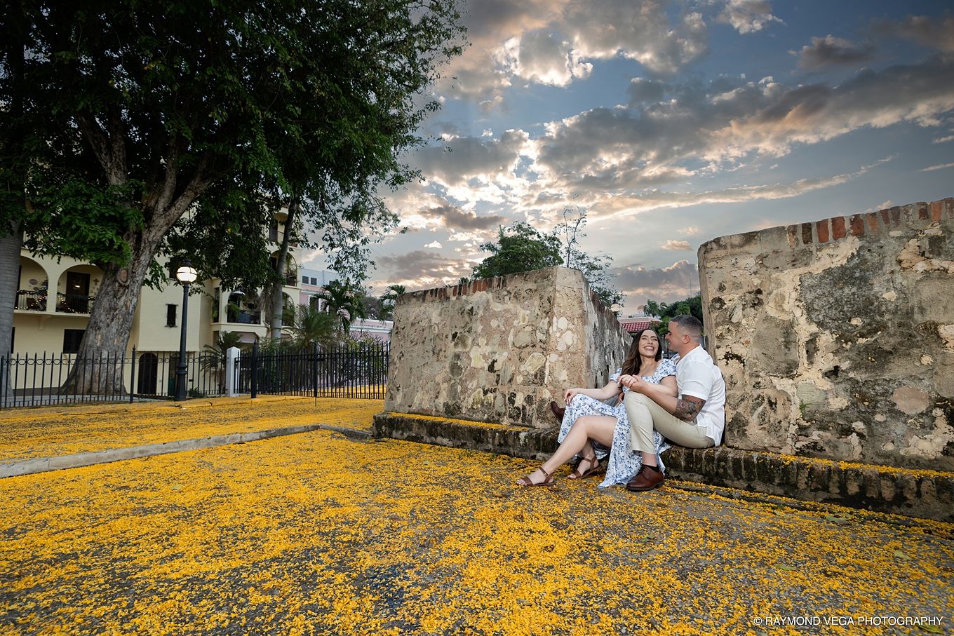 Couple sitting on steps surrounded by yellow flowers with ancient stone walls and dramatic sunset sky in historic city. - Love Story
