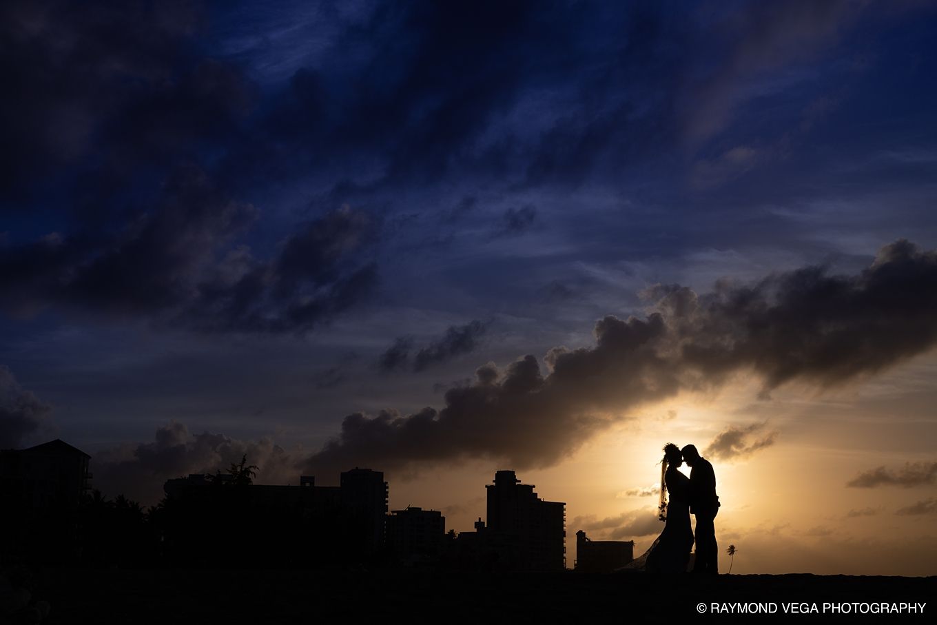Romantic couple silhouette kissing at sunset with dramatic cloudy sky and city skyline backdrop.