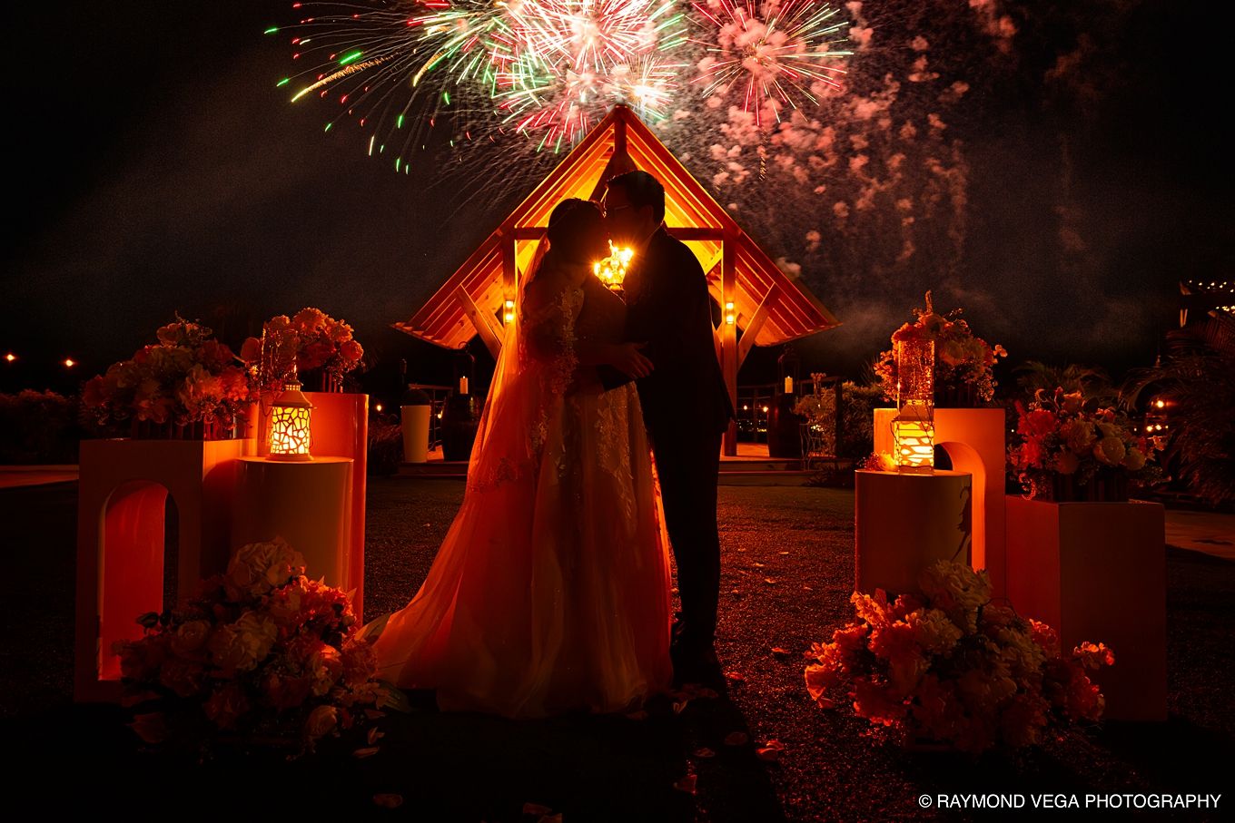 Bride and groom silhouetted against fireworks display at night outside a chapel with floral arrangements.