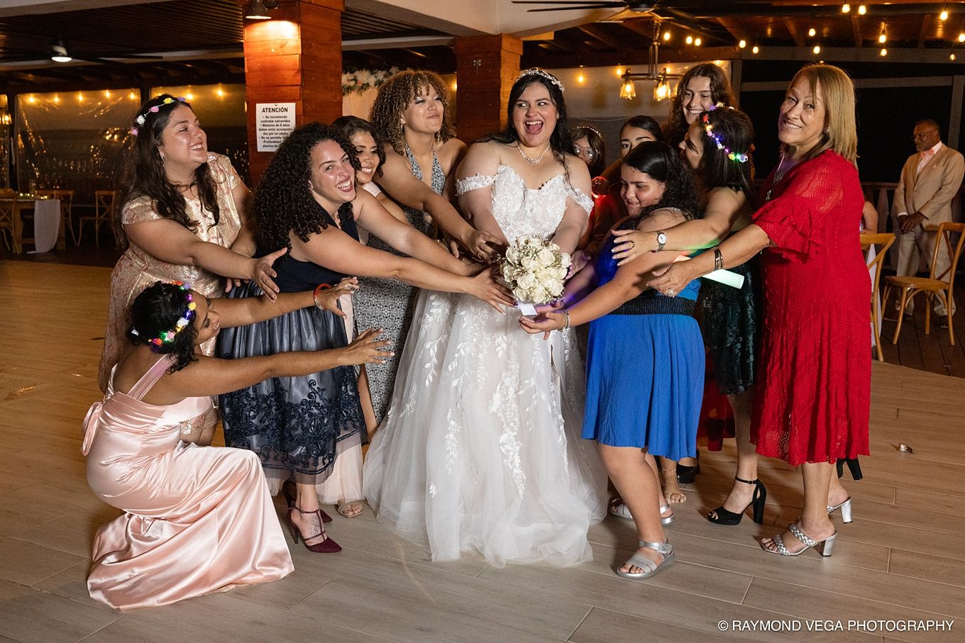 Bride in white gown poses with bridesmaids reaching for bouquet at elegant wedding reception venue.